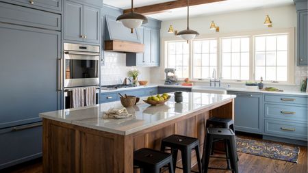 A transitional kitchen with blue-gray cabinets, a wooden island with white countertops, black bar stools, and wood ceiling beams. Brass wall sconces by the window and a butler's sink. 