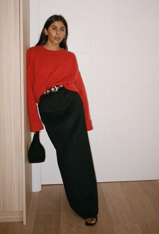 a black maxi skirt outfit showcasing a woman wearing a red crewneck sweater styled with a black belt with silver studs, black maxi column skirt, black heeled sandals, and a black handbag