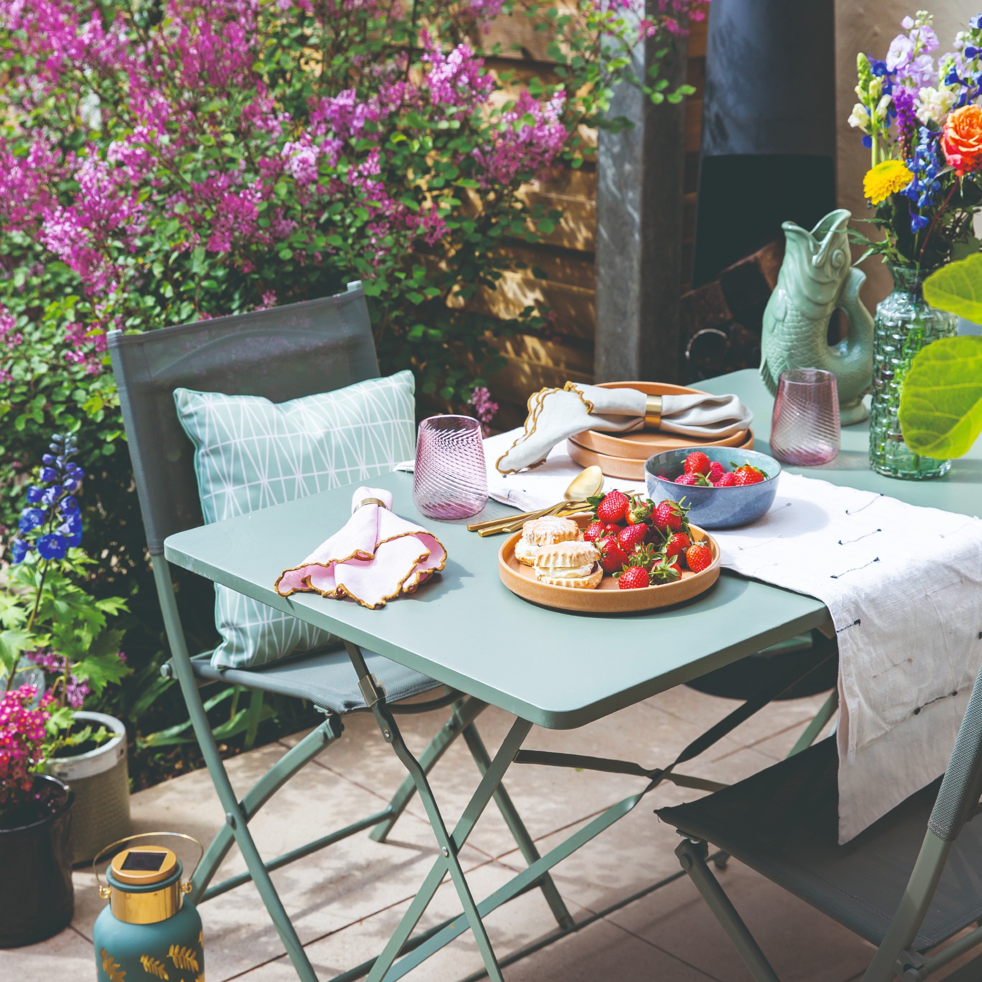 A sage green bistro set on a patio with a vase of flowers and a fish glug jug