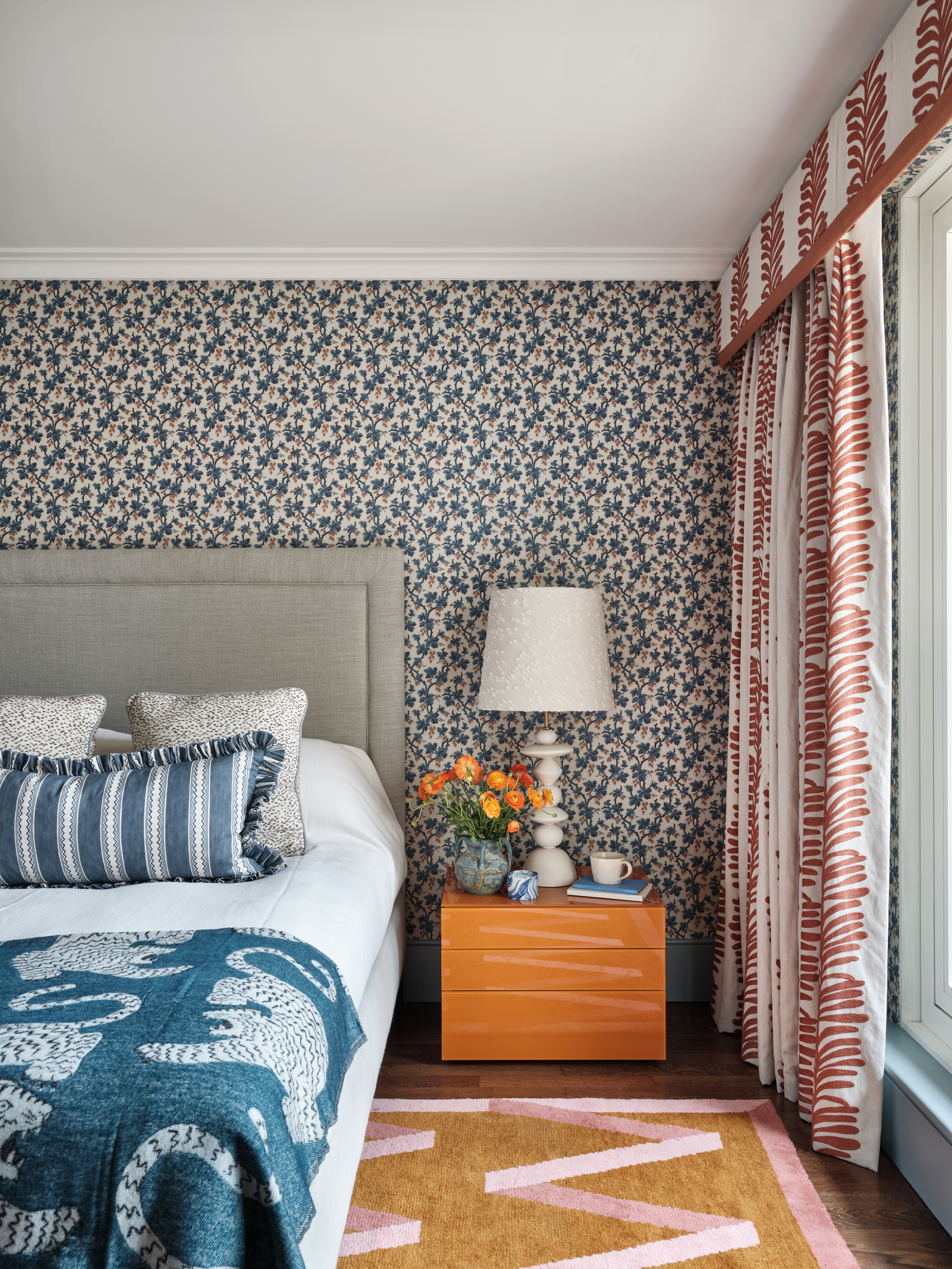 A bedroom with blue floral wallpaper, red and white patterned curtains, and white bedding with a blue patterned throw and lumbar pillow