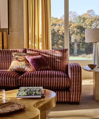 Bright living room with tall windows in background. Red striped upholstered sofa with wavy wooden coffee table. Furnishing such as mustard yellow curtains, burgundy and yellow throws, and neutral beige lamp create a cozy look