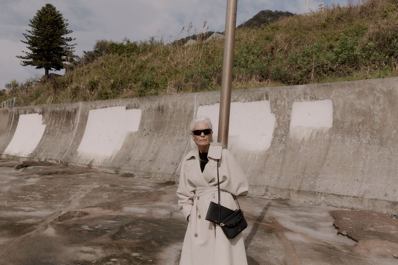 Older lady holding under-the-radar bag label Yu Mei bag and sunglasses against concrete background