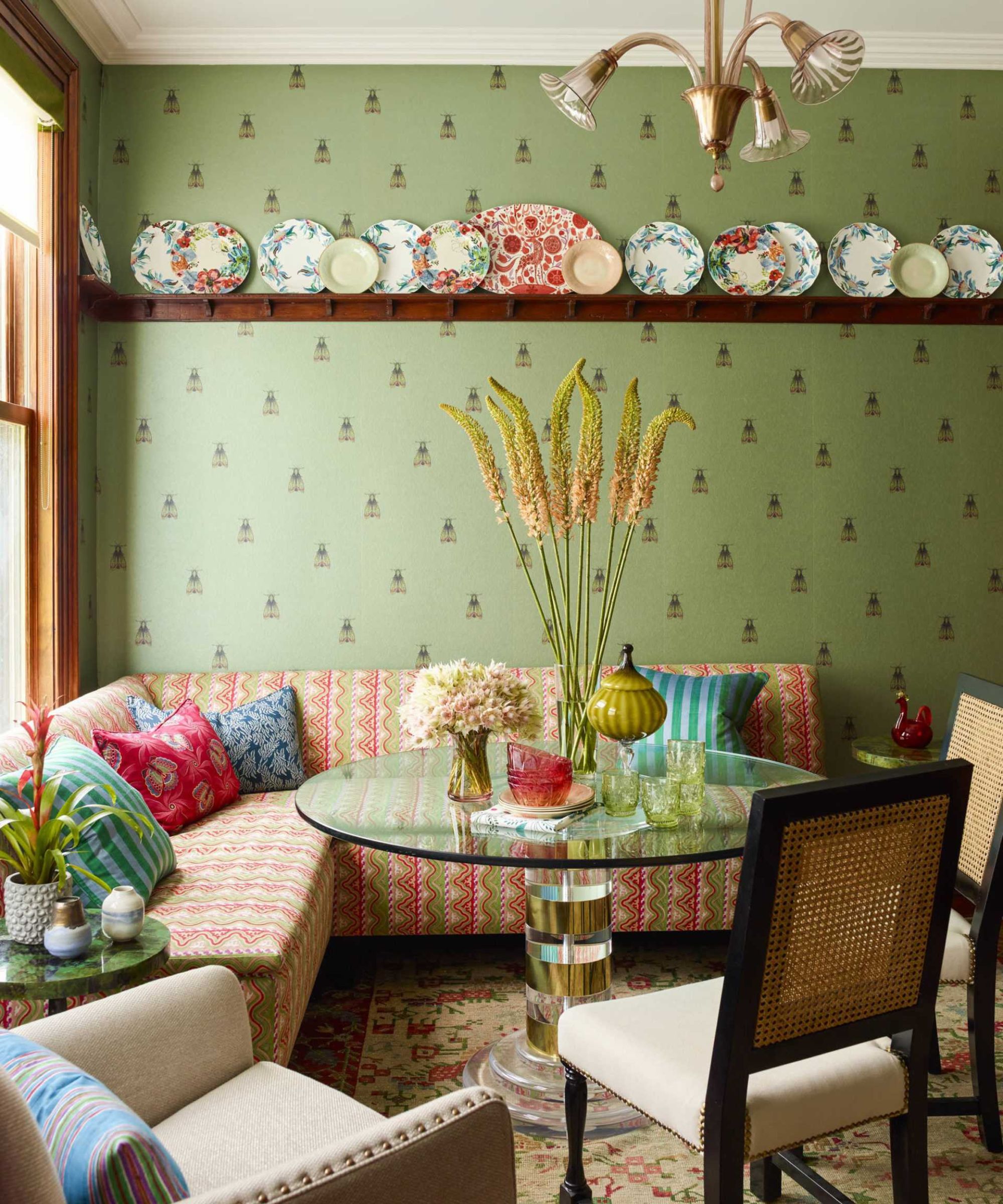 A vibrant dining nook with sage green bee-patterned wallpaper, a colorful patterned banquette, and a glass-top table. A wooden plate rail displays decorative ceramic plates above the seating.