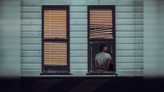 A woman facing away looks out from an open window with blinds, amidst two closed ones on a blue wall