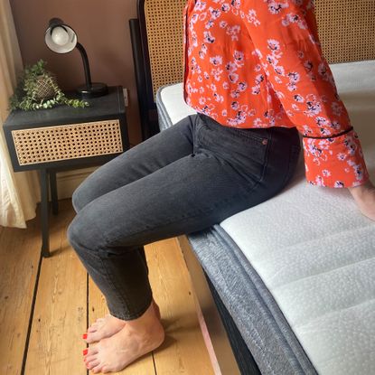 A woman wearing a red floral top and black jeans sitting on the edge of a mattress in a room with wooden floor boards