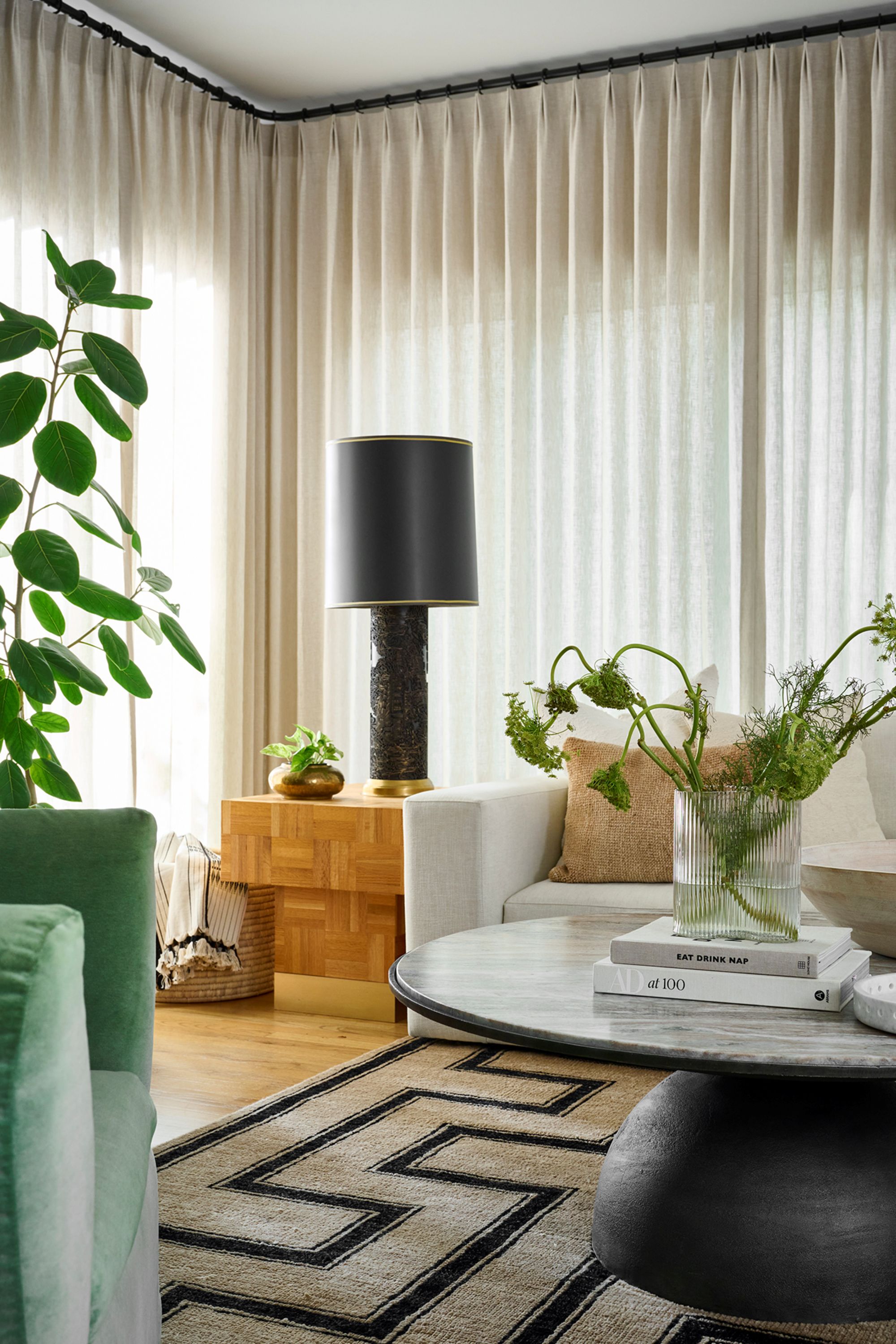 a living room with floor to ceiling curtains, a geometric rug, a large coffee table, and a large black table lamp