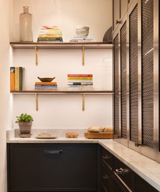 A narrow pantry with dark cabinetry featuring antique brass mesh inserts. Open wooden shelves with brass brackets display stacked cookbooks, bowls, and decorative objects. A white marble countertop holds a potted herb, eggs, and a loaf of bread. Soft lighting highlights the space, casting a glow on the neutral walls.