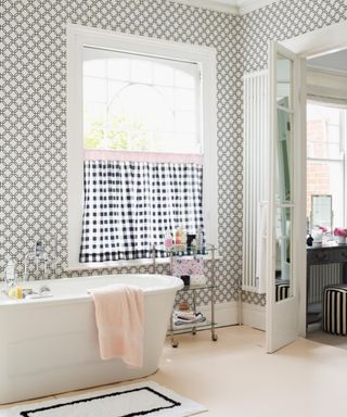 A bathroom with black and white wallpaper, a window with a black and white checked curtain, a white bathtub with a pink towel, a white and black bath mat, beige flooring, and an open door