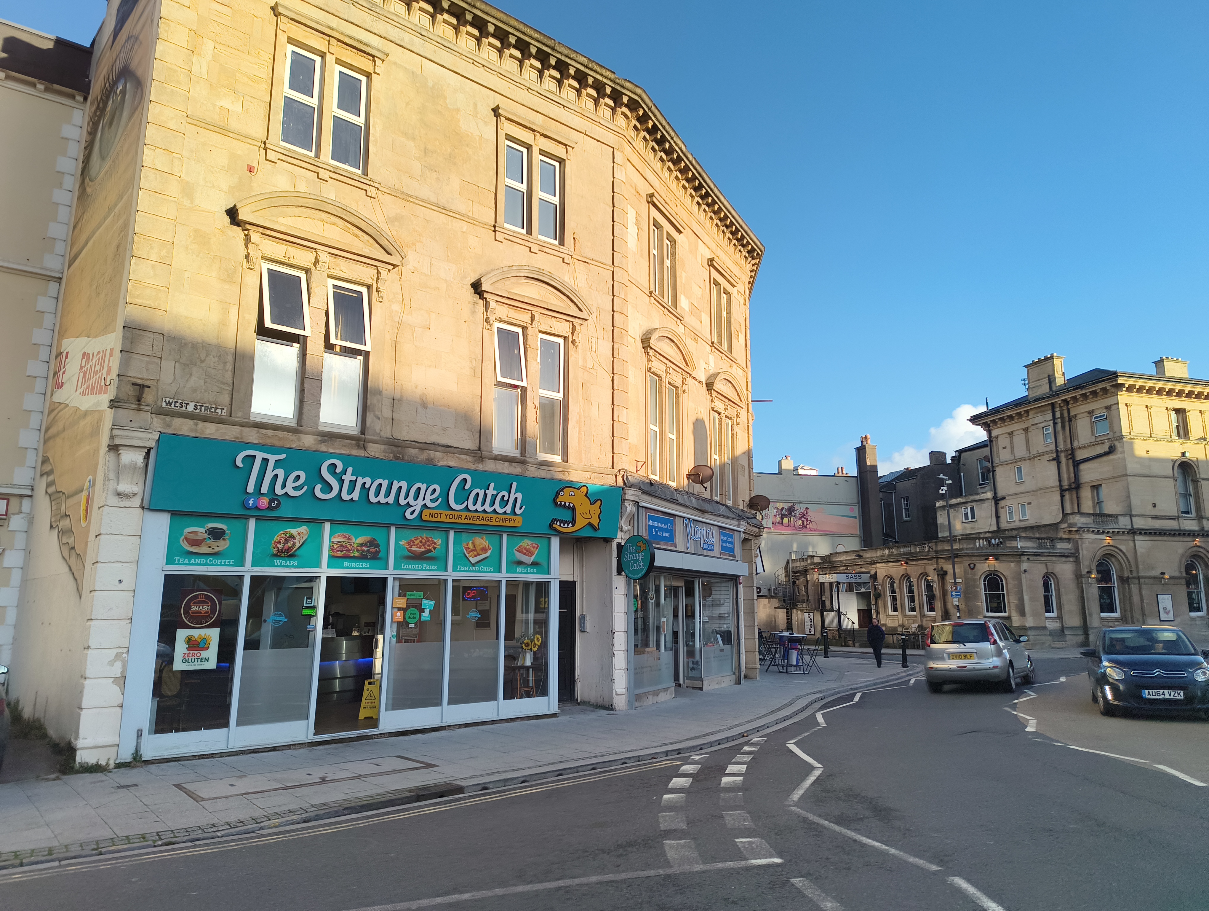 A fish and chip shop named "The Strange Catch" with bright teal signage is located on the ground floor of a yellow-stone historic building on a street corner.