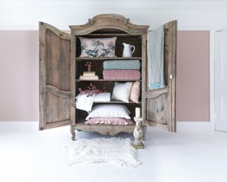 Pretty French provincial style boudoir with rustic wood armoire, displaying open doors and shelves styled with pastel quilts and tactile pillows.