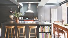 A black kitchen with a silver metal island with brown wooden stools, next to an in-built bookshelf filled with recipe books. On the top of the island is a copper jug filled with green leafy steams, and a silver faucet. Behind is a red hob and silver extractor fan above. To the right is a wooden dining table with matching chairs.
