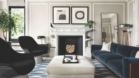 Living room with pale grey walls, blue velvet sofa, two black armchairs, a pale grey ottoman, a geometric blue rug, a large white fireplace and modern art hanging above it.