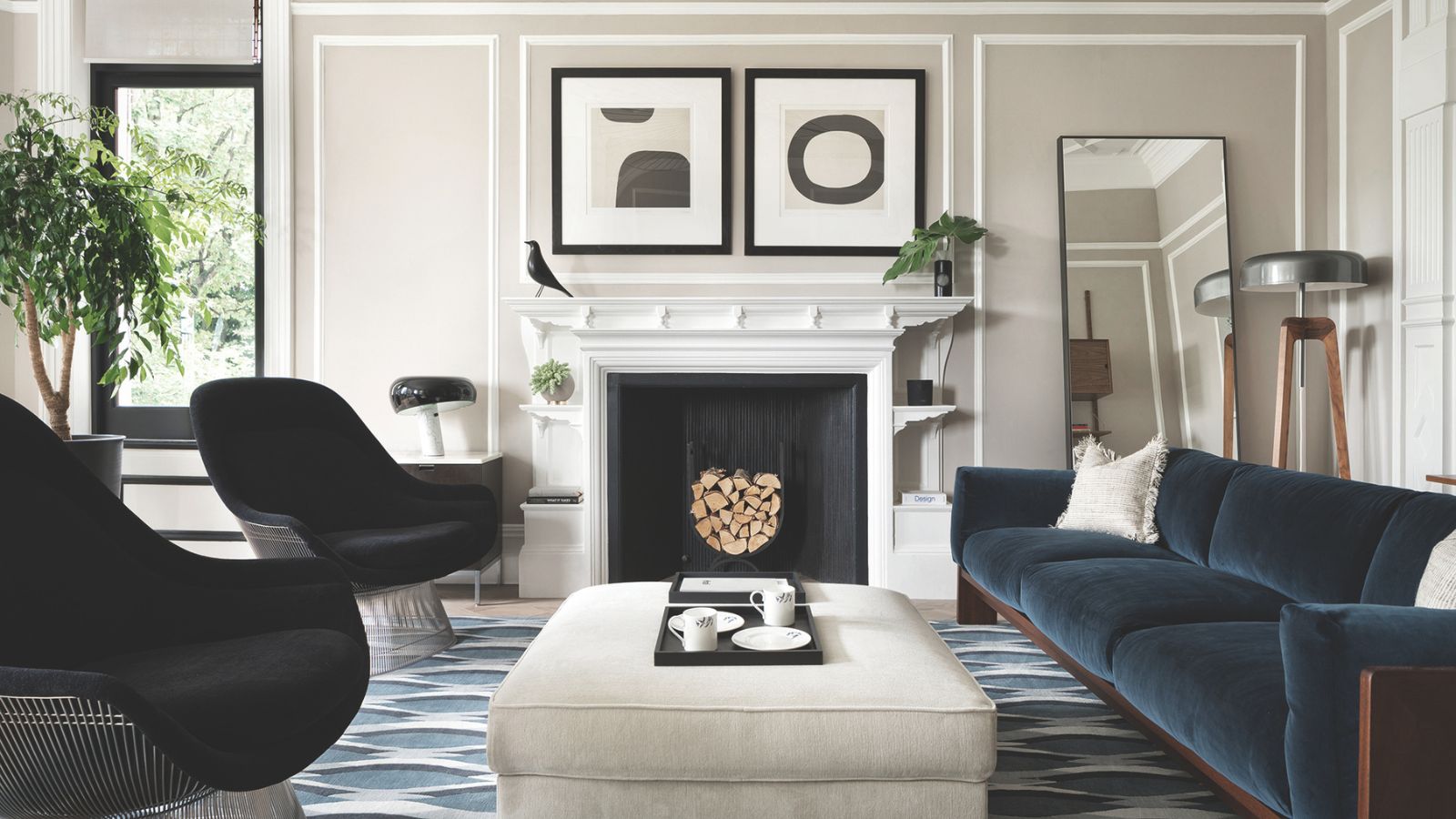 Living room with pale grey walls, blue velvet sofa, two black armchairs, a pale grey ottoman, a geometric blue rug, a large white fireplace and modern art hanging above it.