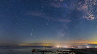 Mostly green Perseids appear over the sea. A faint Milky Way galaxy core and a few light clouds can be seen as well. Light pollution in the distance.