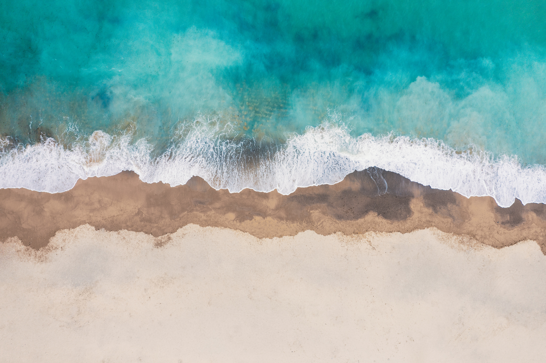 A view of a beach from above, showing the sea and the sand. The sand is very white, the sea is very blue