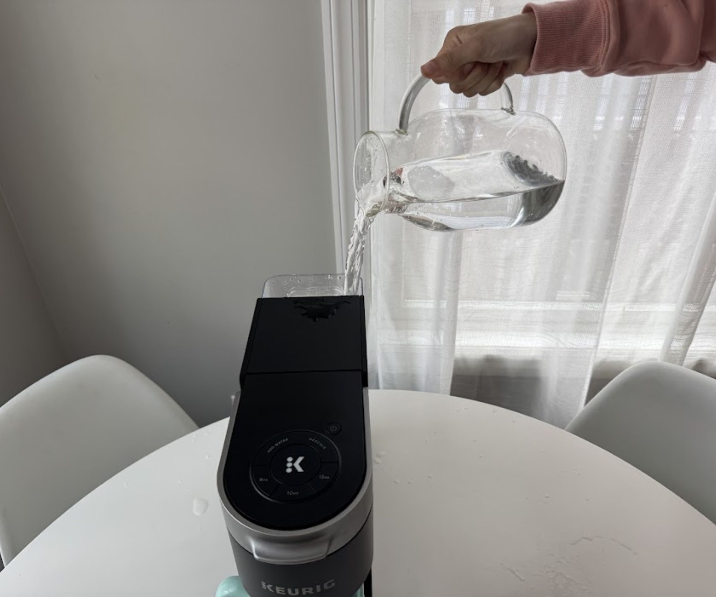 A hand pouring water from a glass pitcher jug into the reservoir of a black Keurig coffee machine.