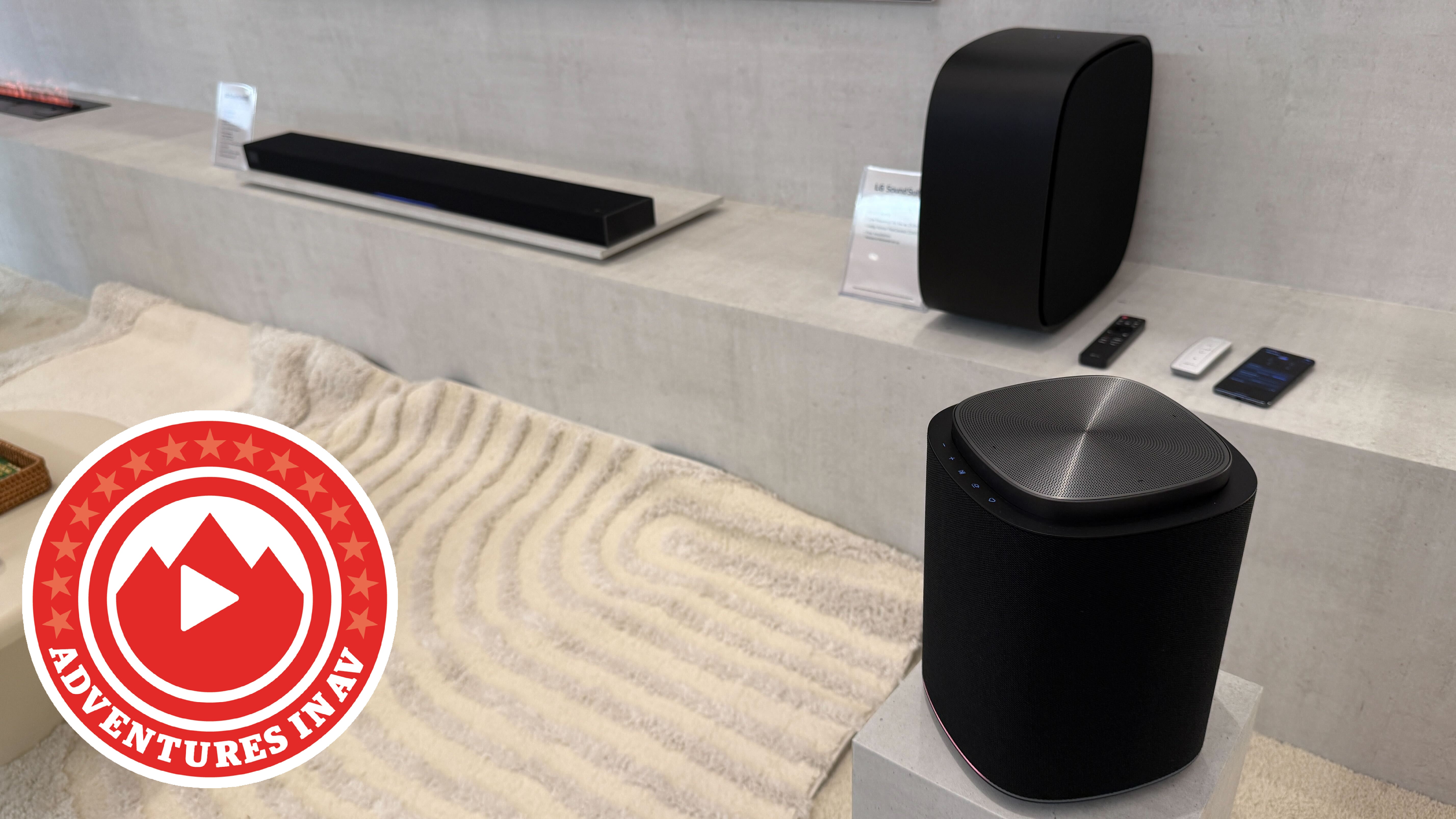 The LG Sound Suite H7 soundbar, M7 wireless speaker, and W7 subwoofer in a modern concrete-style room with a textured rug on the ground