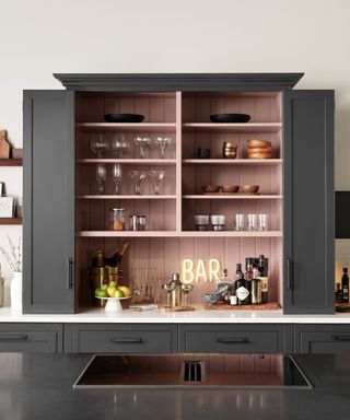 An open black kitchen cabinet with dusky pink shelves with storage jars and mugs in it, with a black stovetop in front of it