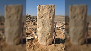 Here we see a rectangular stone standing upright with carving on it in the middle of a desert.