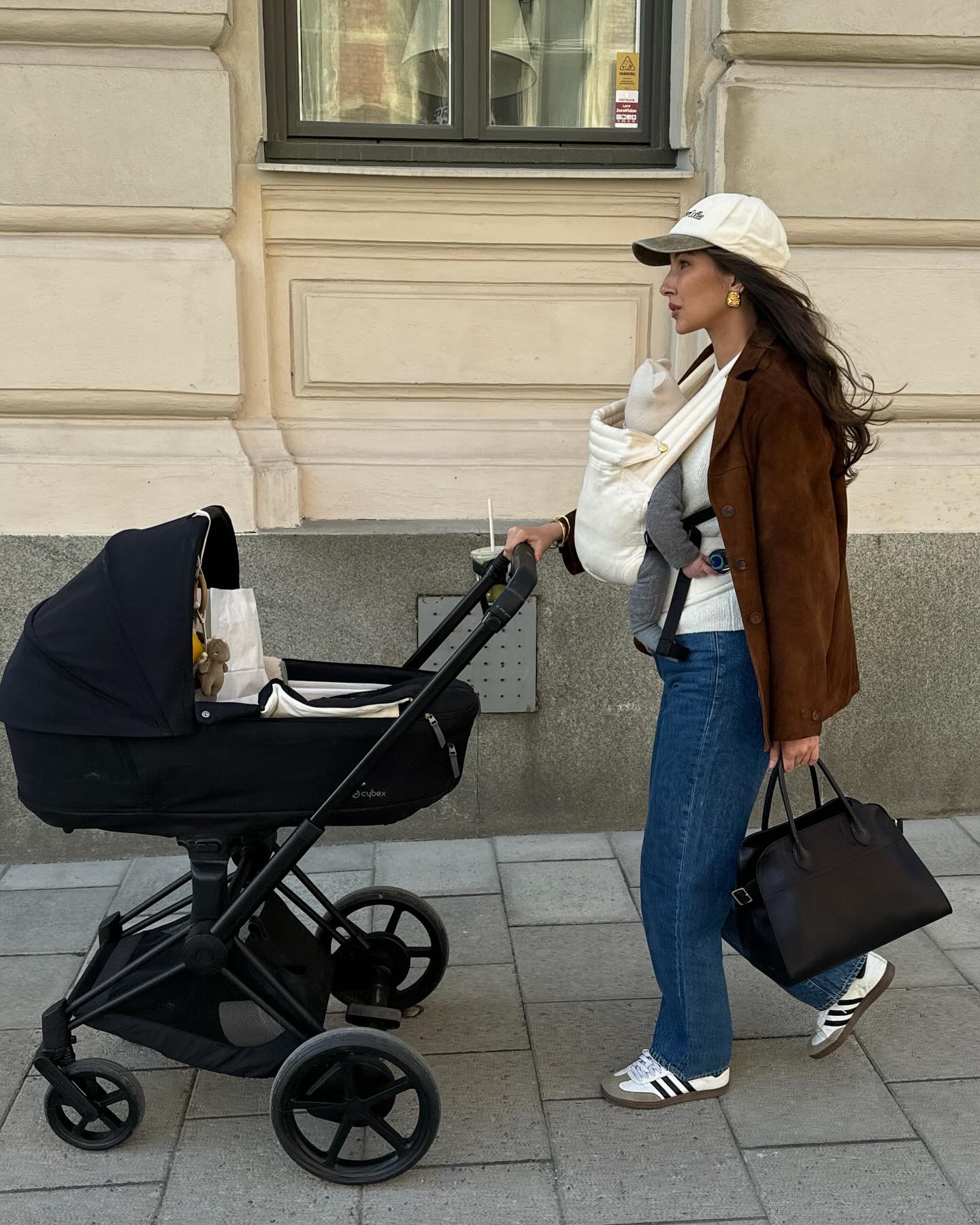 A new mom carries her baby while wearing a brown suede jacket with jeans and Adidas sneakers.