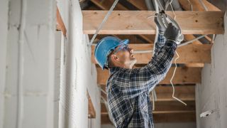 A man in a blue hard hat working on wiring in a roof void