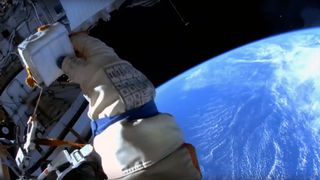 A cosmonaut's gloved hand is seen against the Earth below as viewed from a spacesuit helmet-mounted camera during a spacewalk.