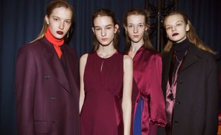 Four female models wearing looks from Paul Smith's collection. One model is wearing an orange turtle neck and dark maroon jacket. Another model is wearing a sleeveless maroon dress. Next to her is a model wearing a maroon top and blue bottoms. And the fourth model is wearing a black turtle neck, necklace, maroon jacket and black coat over the top