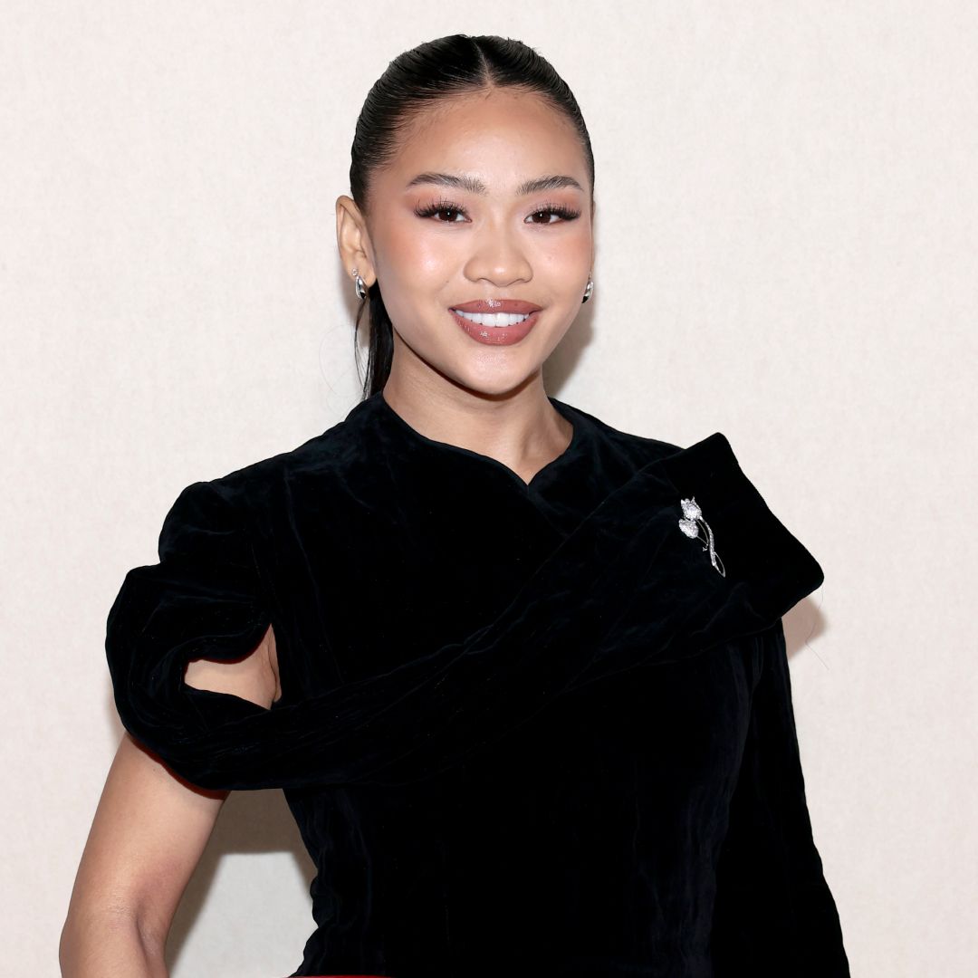 Gymnast Suni Lee wearing a ponytail hairstyle, soft skinlike makeup and a black top. She is smiling at the camera