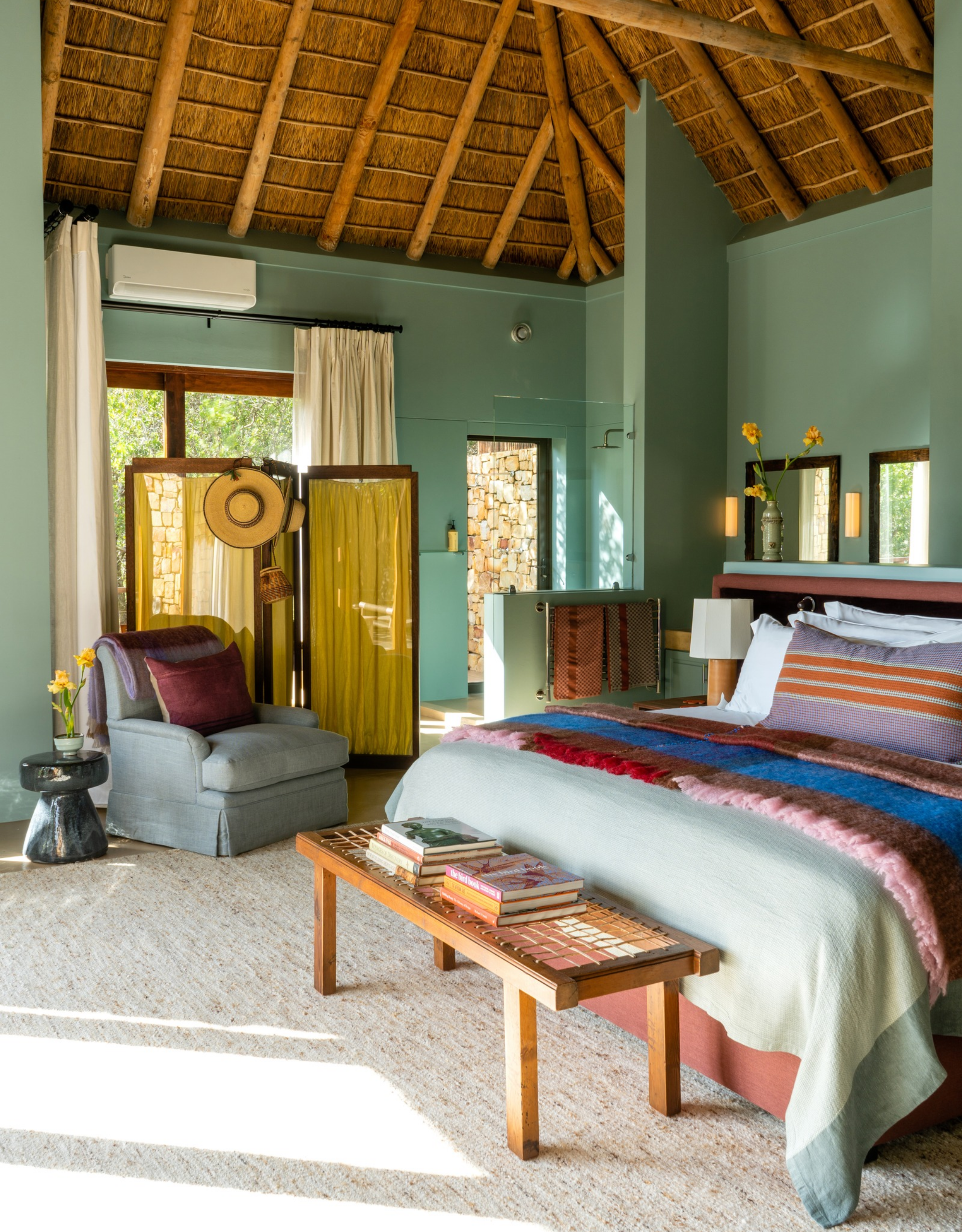 A teal bedroom at the Tamodi Estate with a yellow room divider, a blue chair, a bench, a bed with striped and tufted textiles, and a thatched roof above
