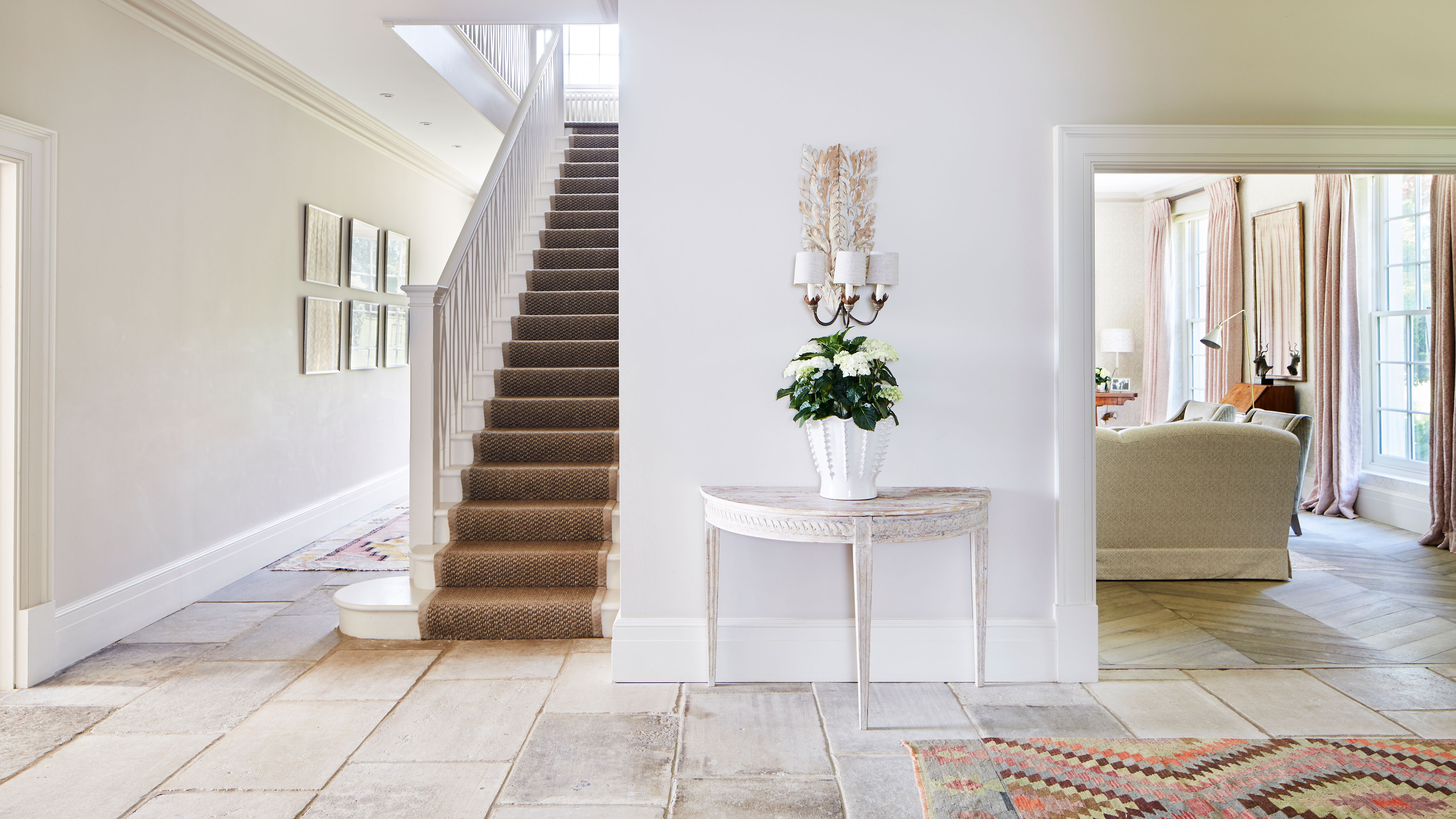 Minimalist entryway design featuring a white console table with fresh flowers, stone flooring, and a classic staircase. The bright, airy interior showcases hidden storage potential and a clean aesthetic.