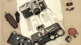 A flat lay photo displays three vintage Leica cameras, two with lenses attached and one bottom-up, surrounded by small accessories and resting on a partially visible poster with the word "Leica."