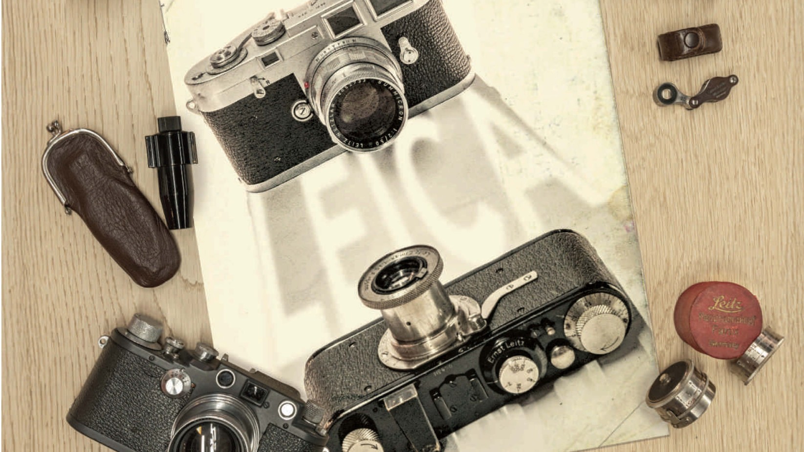 A flat lay photo displays three vintage Leica cameras, two with lenses attached and one bottom-up, surrounded by small accessories and resting on a partially visible poster with the word "Leica."