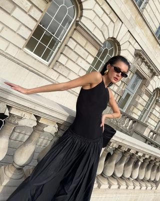Fashion influencer poses on a sidewalk of London wearing large drop earrings, oval sunglasses, a mini black bag, and a black mixed-media dress.