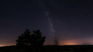 Two meteors are pictured racing through a starry night sky parallel to the Milky Way. Light pollution is visible above a silhouetted horizon and trees.