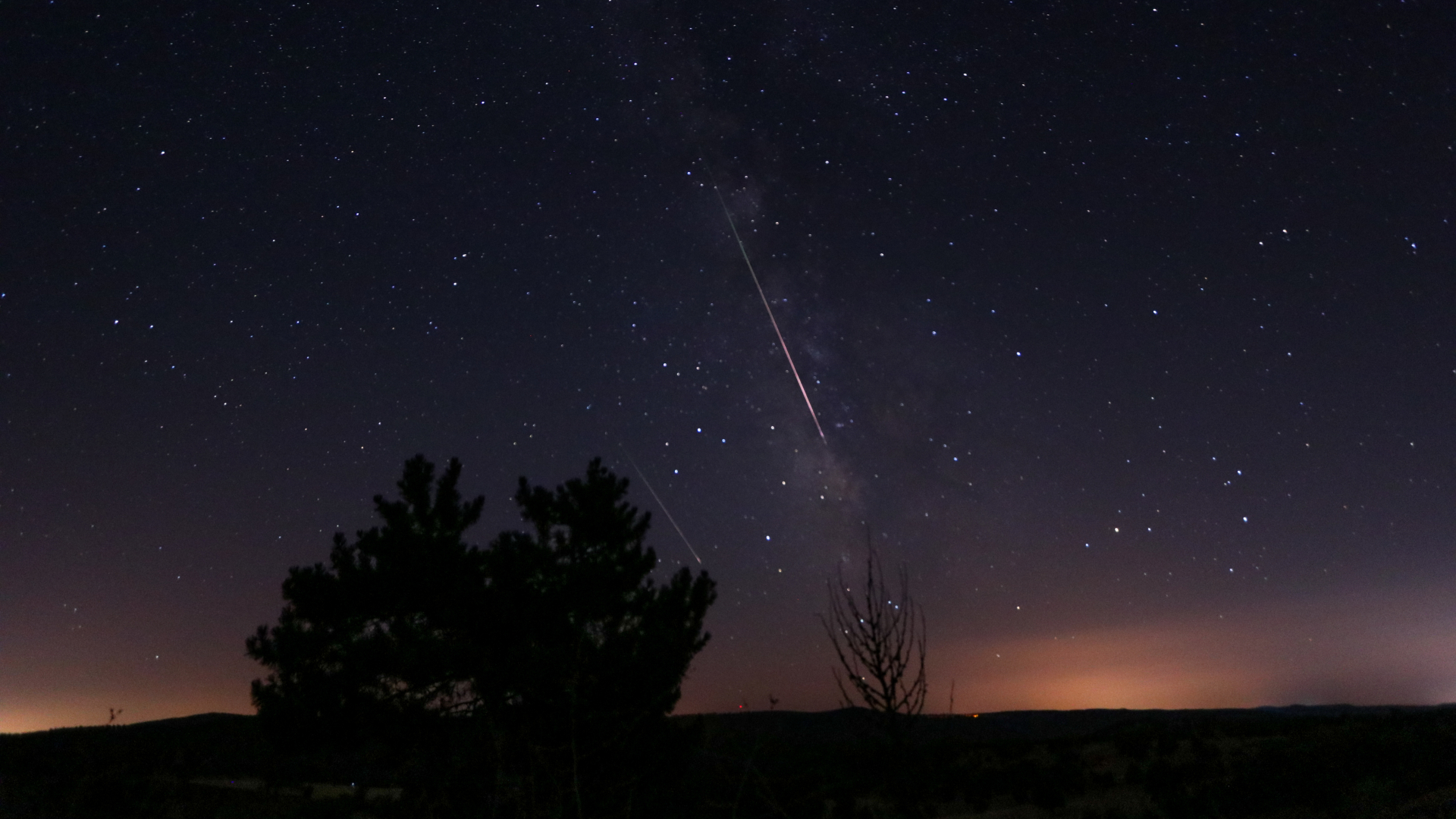 Two meteors are pictured racing through a starry night sky parallel to the Milky Way. Light pollution is visible above a silhouetted horizon and trees.