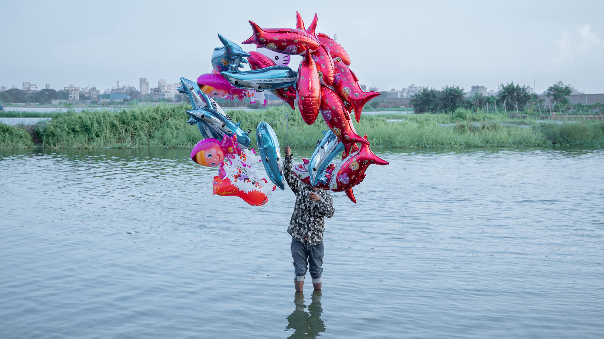 The fish belong in the river, but the river is gone. A man stands in the water, while balloons shaped like dolphins float above, a parody of joy