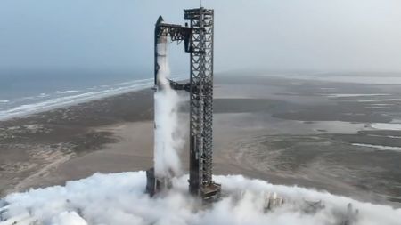 A giant silver Starship rocket is stacked atop its giant booster atop a seaside launch pad for SpaceX's Flight 5. silver SpaceX Starship rocket as it is hoisted atop its booster.