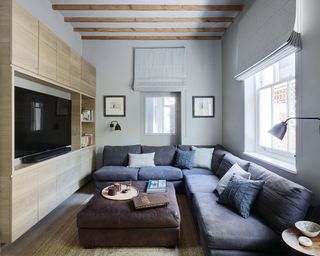 Living room with large gray L shaped sofa, ottoman and built in cupboards with television, wooden beams, gray painted walls and blinds, wall lights and framed artwork, textured rug on wooden flooring