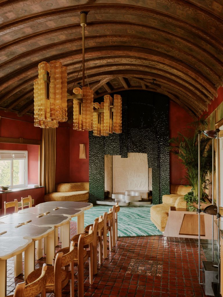 A beautifully designed living room features an organic shaped table with plenty of wooden chairs, Murano glass chandeliers, wooden arched ceilings, and modernism-inspired furniture, all placed on terracotta tiles.