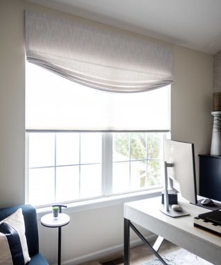 A home office with a large window with white folded blinds at the top on a white wall, with a white desk with a silver computer to the right of it and a blue seat with a pillow on to the left of it