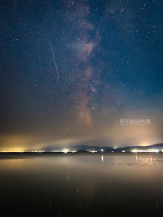 a vertical image shows a long perseid meteor streak in the sky with the milky way bulging in the center and a distant glow of yellow in the clouds.