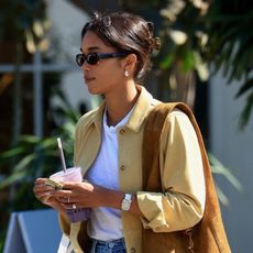 Laura Harrier walking with a smoothie in LA wearing a soft-yellow top, a white tee, vintage Levi's jeans, a brown suede bag, and black slides.