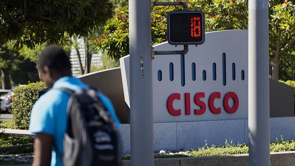 A man walking past traffic lights with a sign in the background displaying the word Cisco