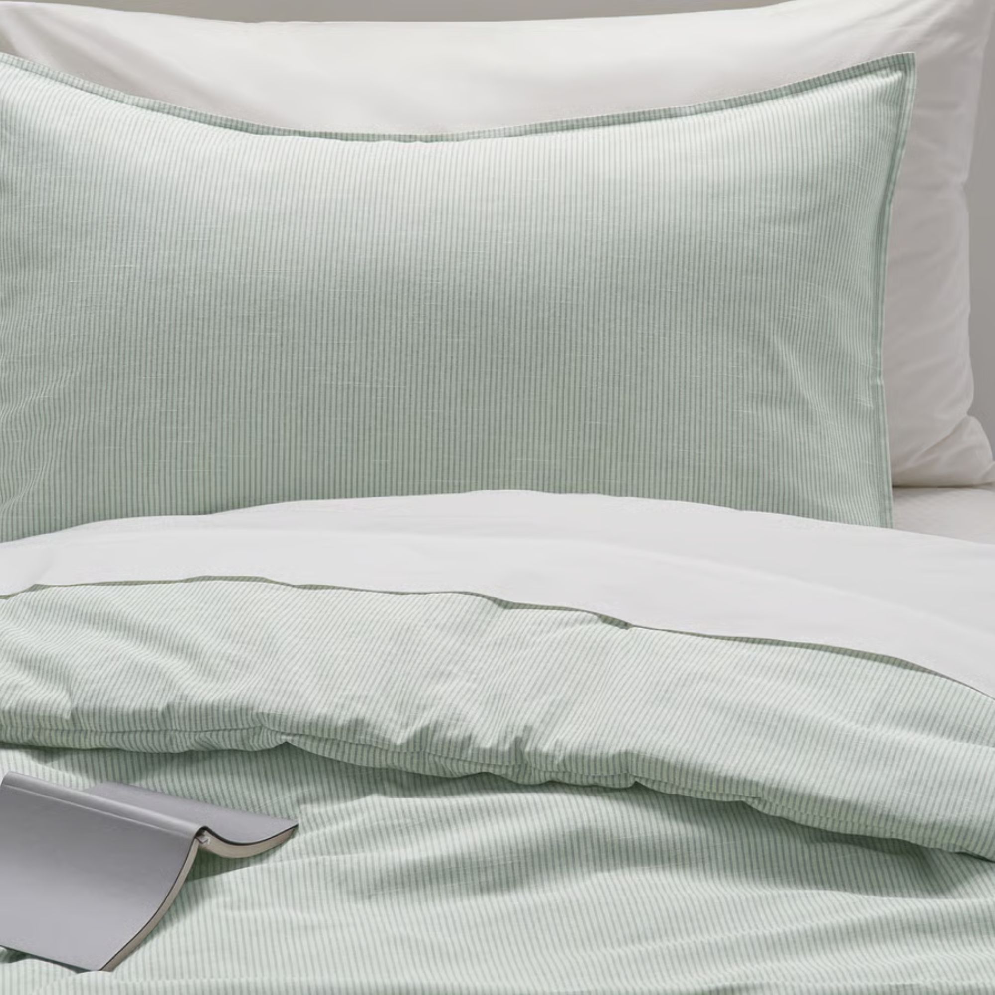 Sage green striped IKEA BERGPALM bedding on a bed with white sheets and a white pillow, and an open grey book on the bed.