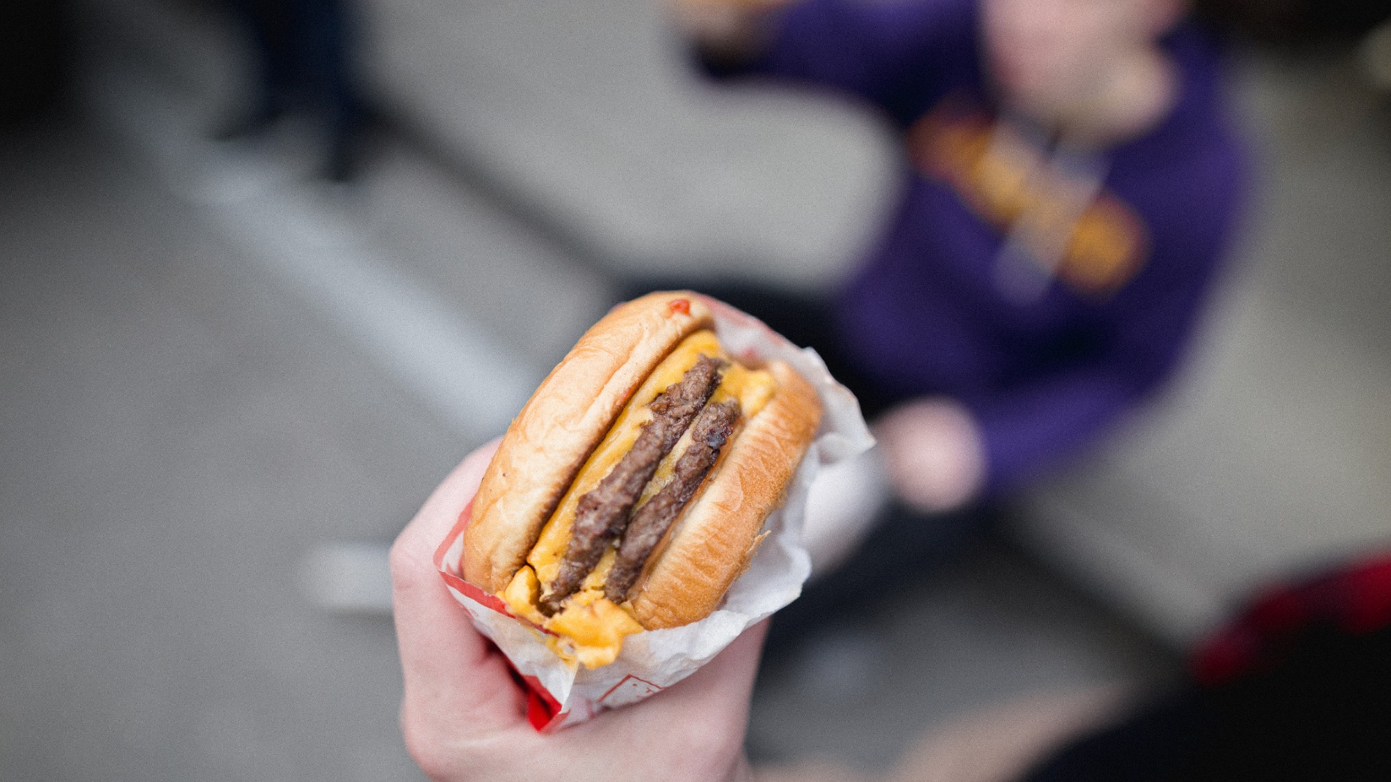 in focus shot of a person holding a fast food burger. in the blurred background is a person seated with a purple hoodie