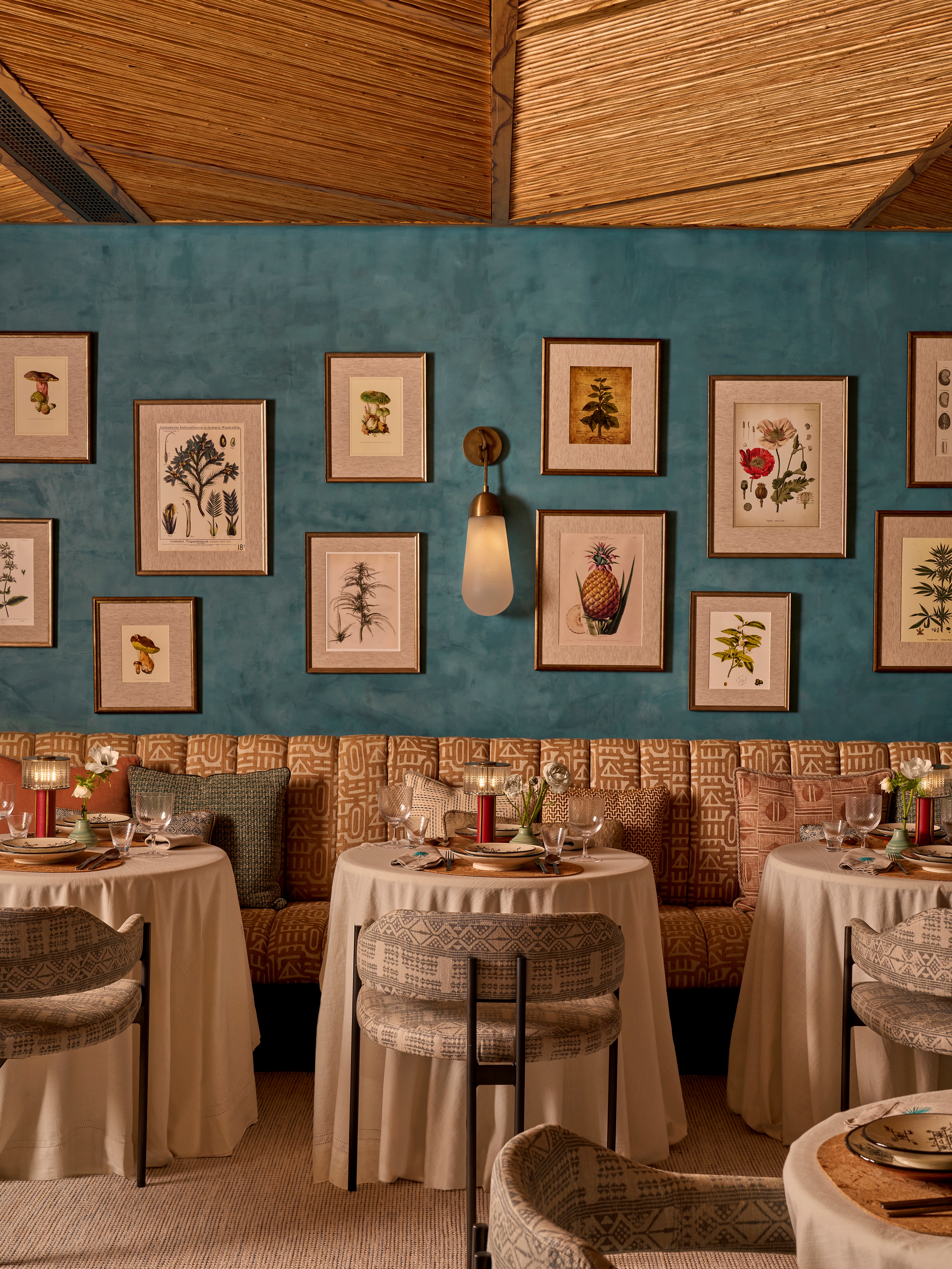 A series of interior shots of a turquoise plaster-tinted restaurant with textural walls and earthy furnishings throughout, from leathery banquettes and cream tablecloths to wooden accents, pendant lighting, and a rattan and wood ceiling.