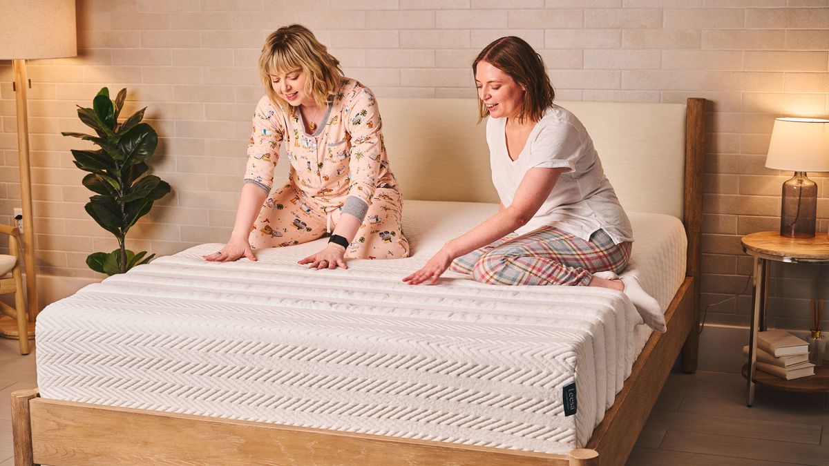 Two women sit on a Leesa Legend Hybrid Mattress in a bedroom, testing the feel of the mattress.