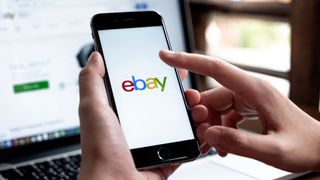 A person holding an iPhone displaying the eBay logo with an eBay listings page displayed on a laptop screen in the background.