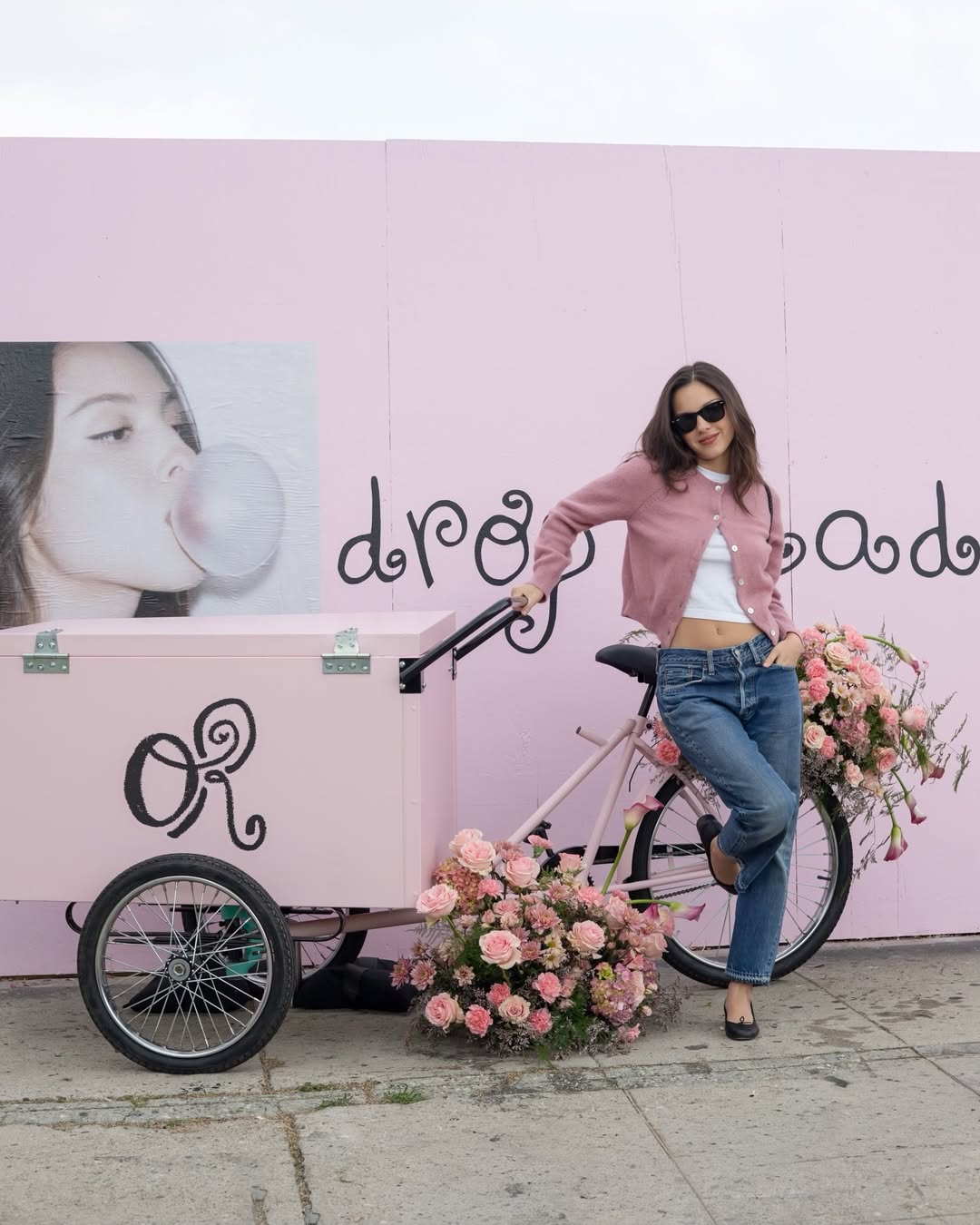 Olivia Rodrigo wears a pink Doen cardigan with blue jeans and a white T-shirt and carries a black woven bag