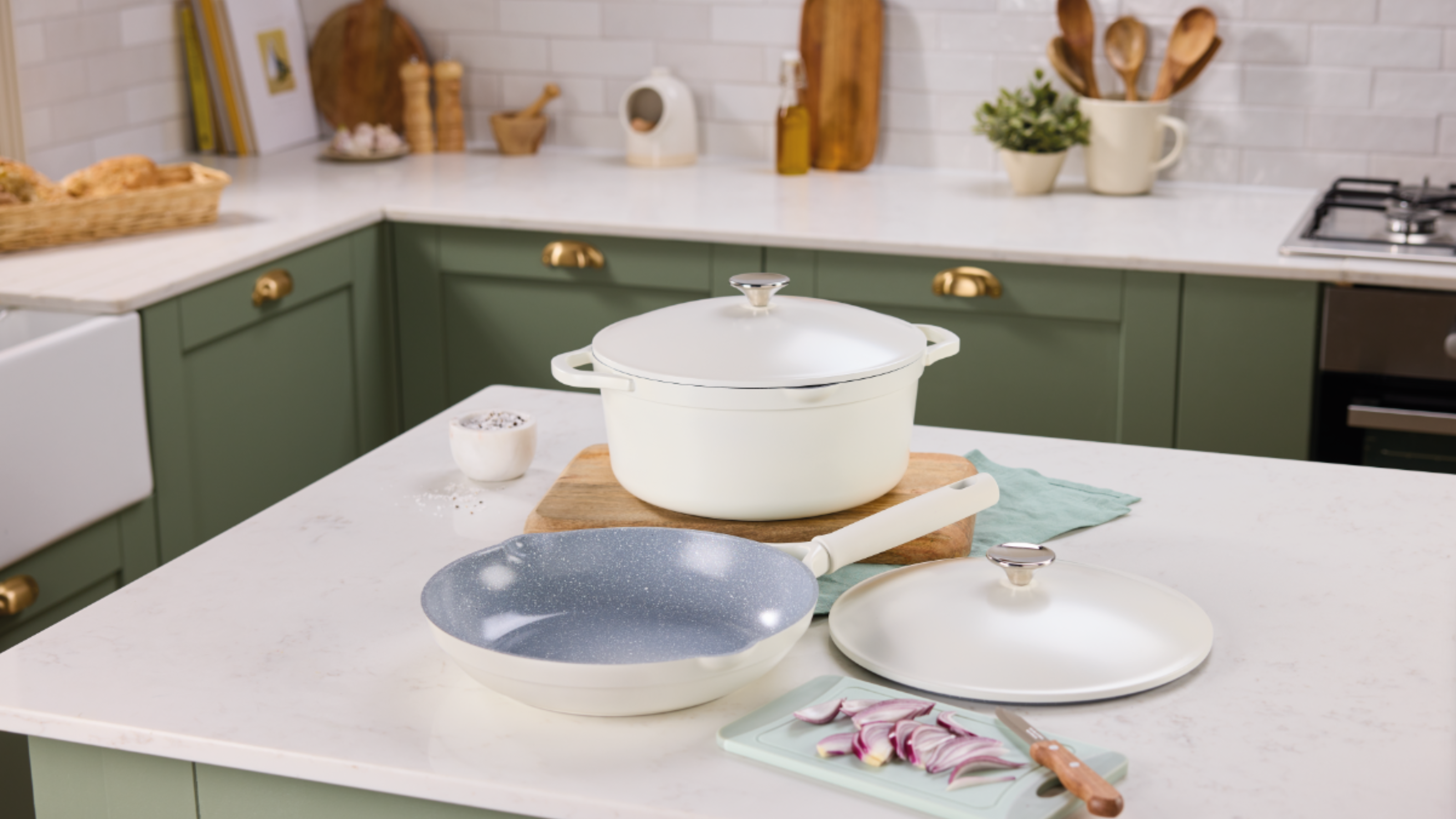 Aldi aluminium frying pan and casserole on a white kitchen island in a kitchen with white countertops and green cupboards.